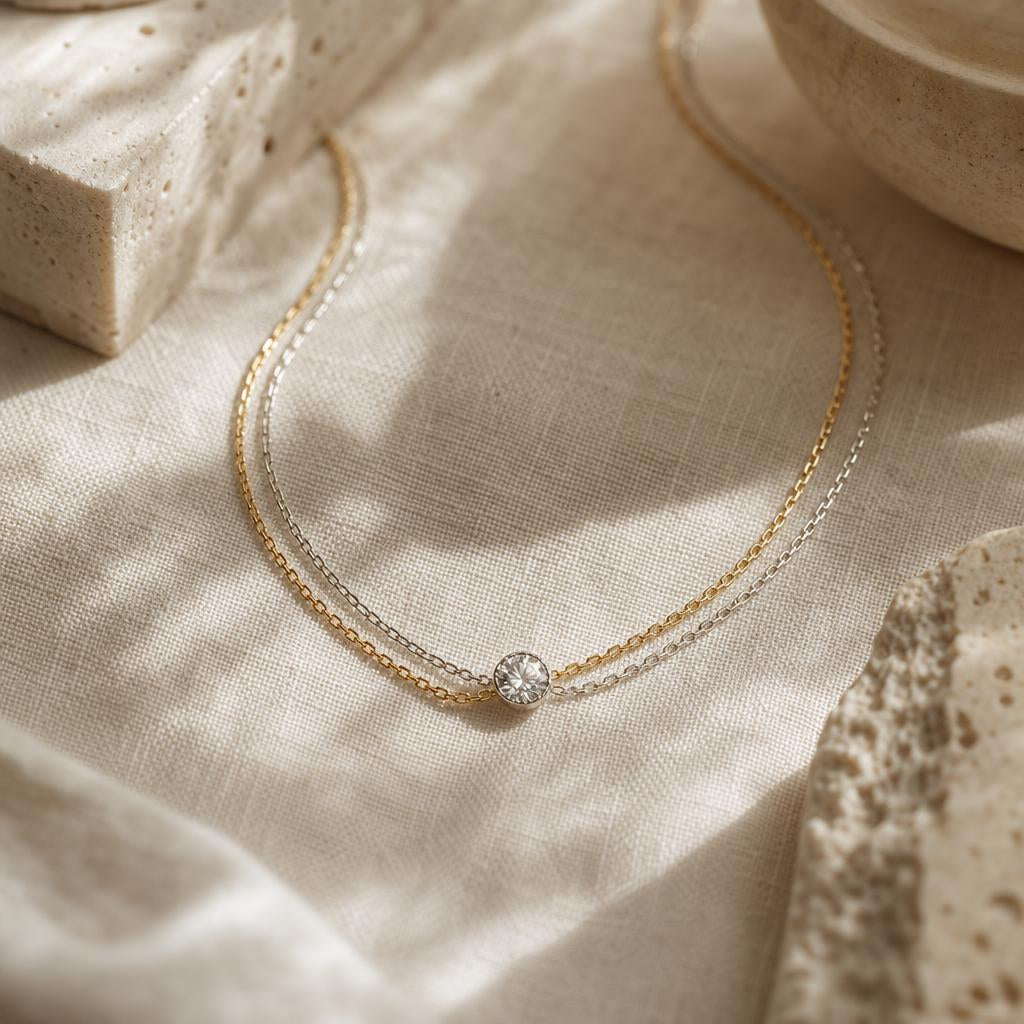 Gold and silver-toned chains curled together on warm cream linen with a sparkling stone