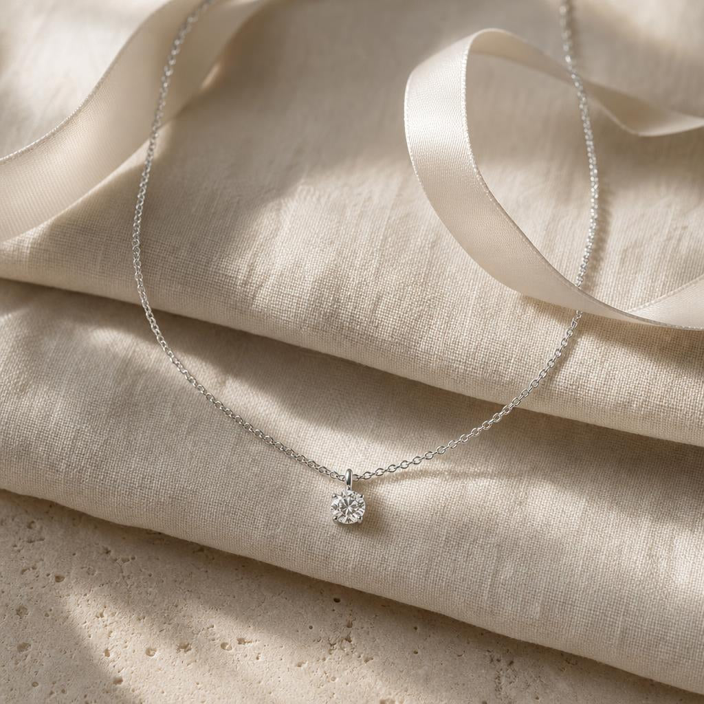 Fine silver chain with a small sparkling pendant beside an ivory ribbon on cream linen