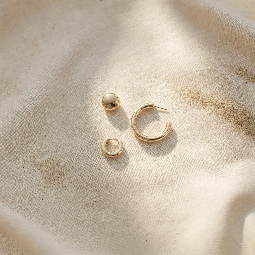 Three earring shapes — stud, hoop, and huggie — styled on warm cream linen