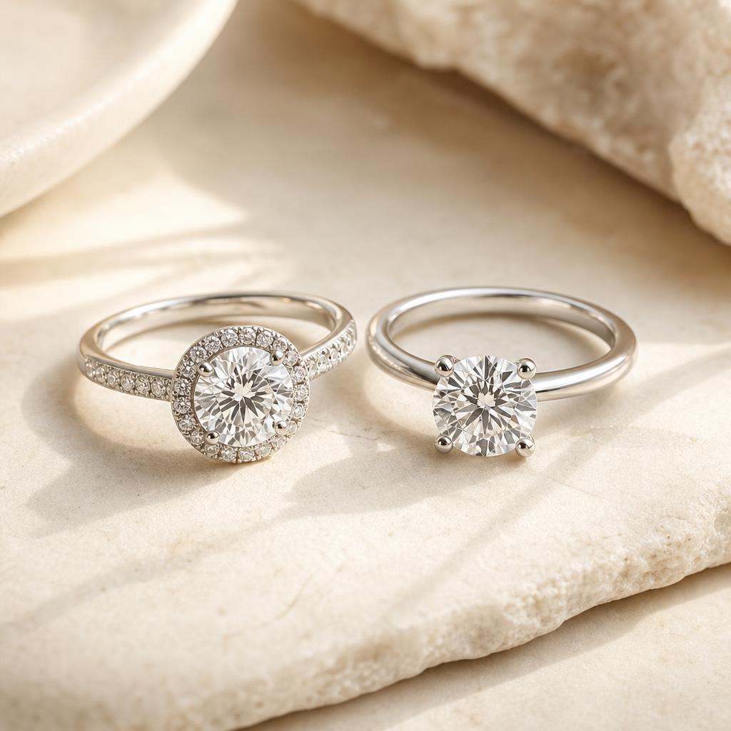 Two engagement rings side by side — one halo, one solitaire — on a warm cream stone surface