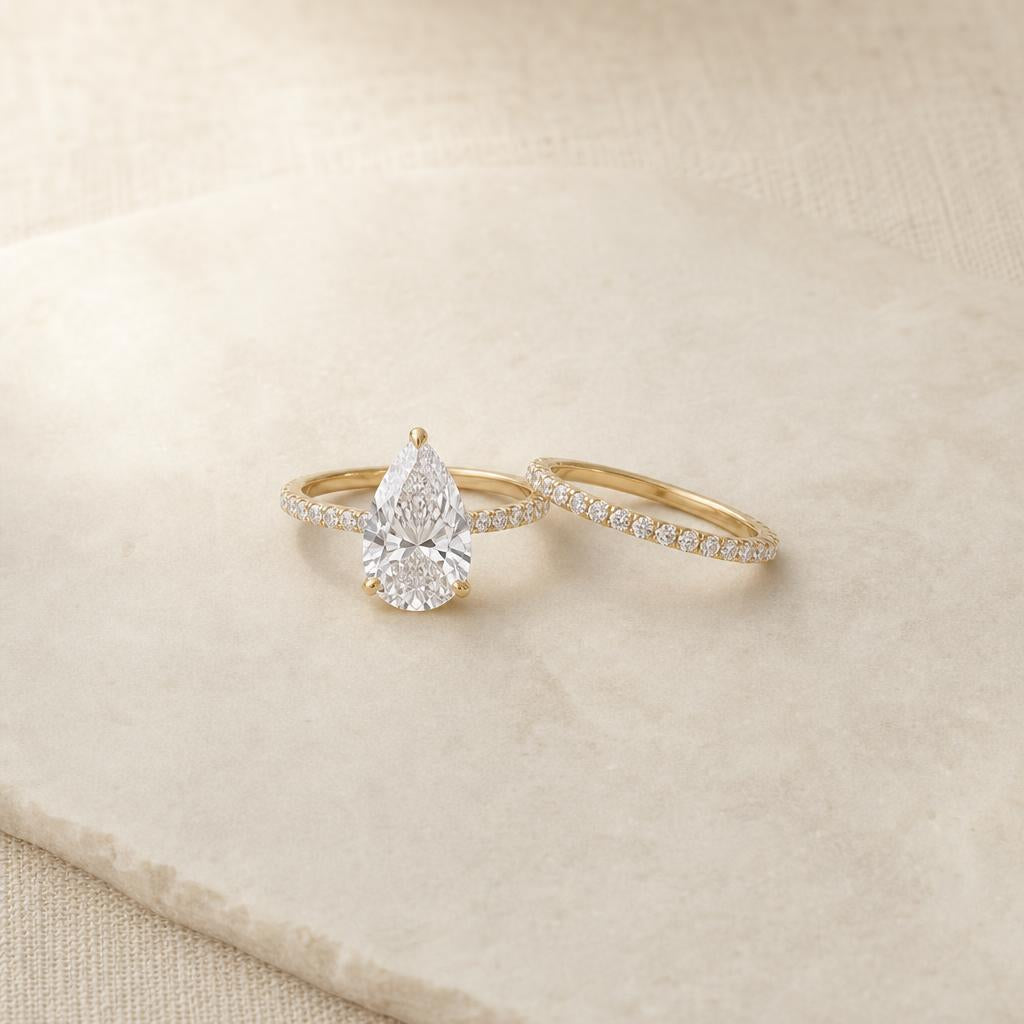 Pear cut engagement ring and slim pavé eternity wedding band on cream stone surface