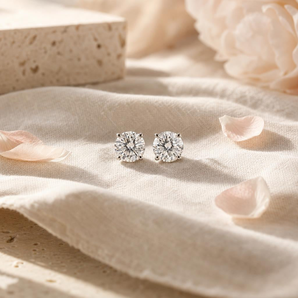 Pair of round diamond stud earrings on cream linen with blush petals