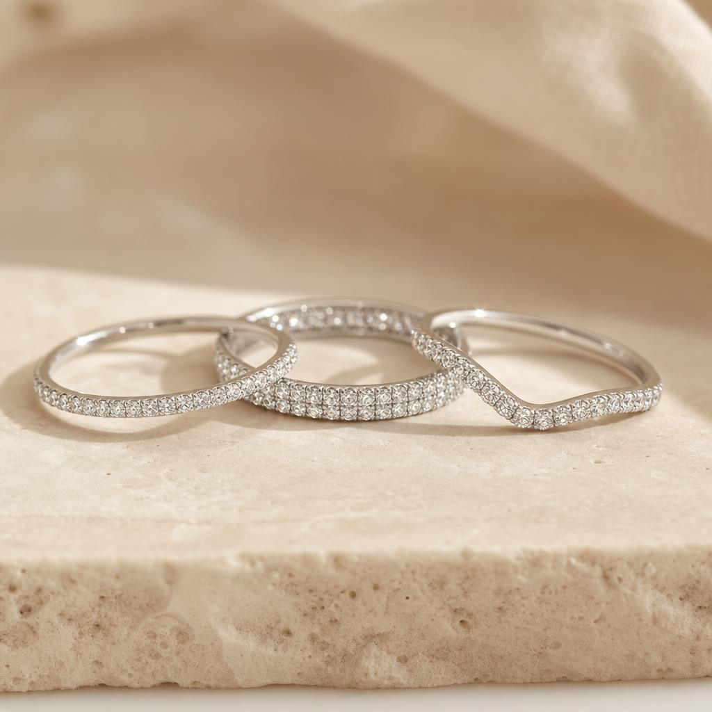 Three diamond stacking bands — slim pavé, bold eternity, and wishbone — arranged on warm stone