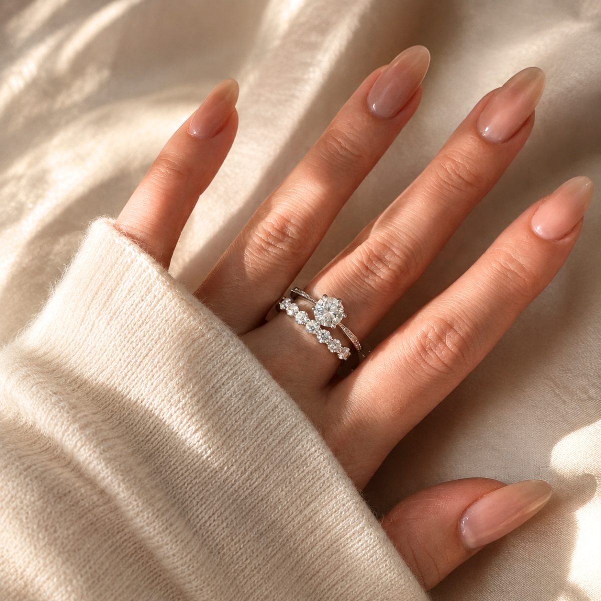 A close-up of a hand wearing a silver engagement ring with a round-cut center stone and a matching band with small pavé-set stones, resting on a soft beige knit sleeve against a draped beige fabric background.